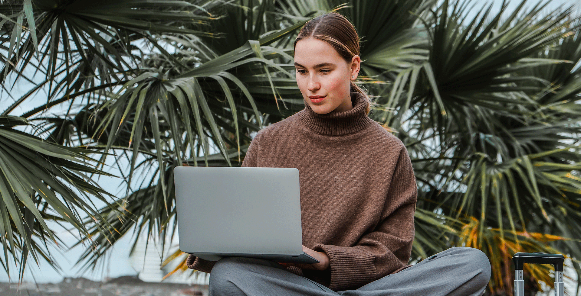 Frau mit Laptop sitzt während einer Workation vor Palmen. © Adobe Stock, shintartanya