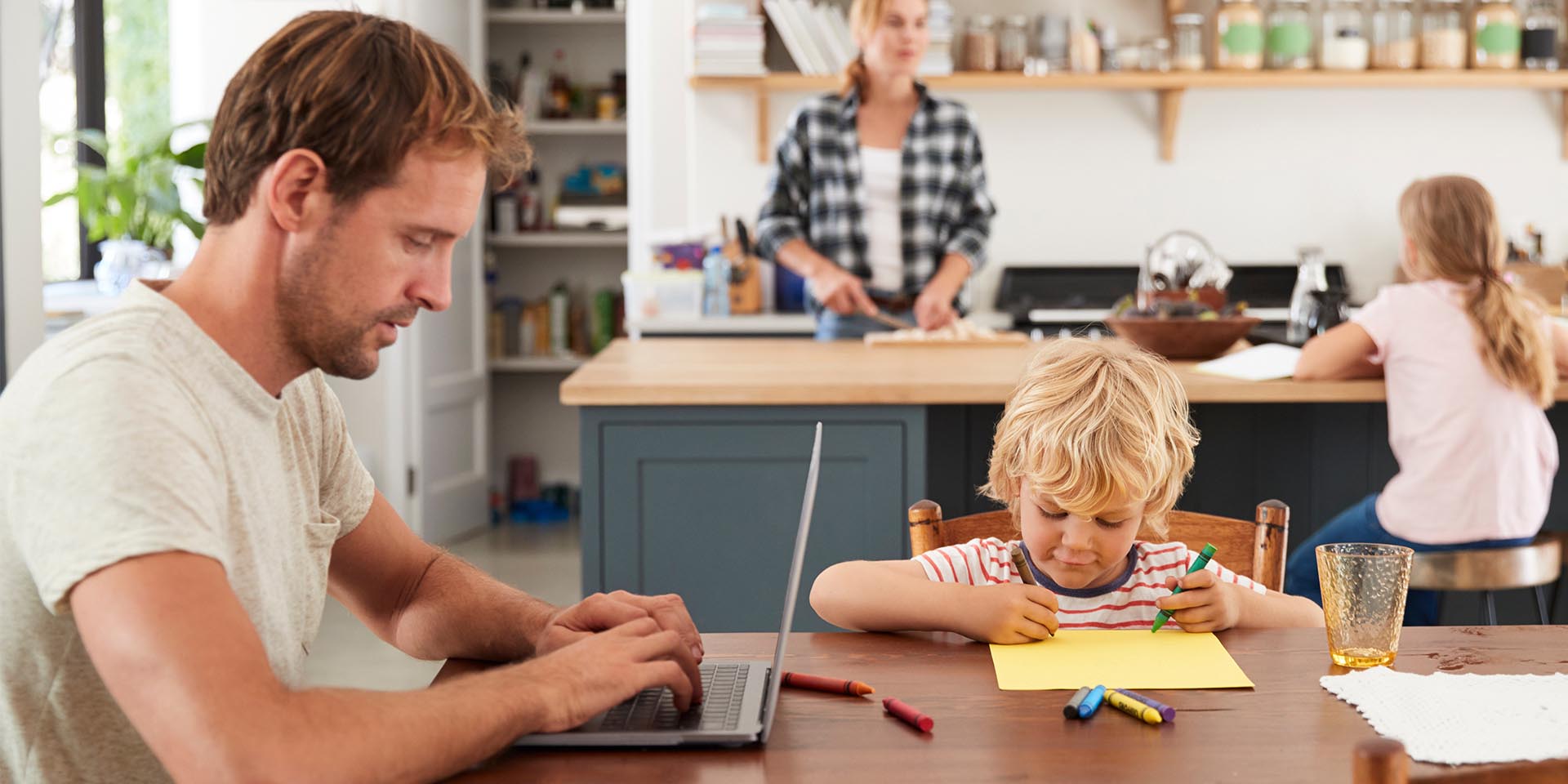 Ein Mann arbeitet von zuhause am Laptop während daneben ein Kleinkind malt und im Hintergrund die Frau kocht und das andere Kind Hausaufgaben macht © Adobe Stock, Monkey Business