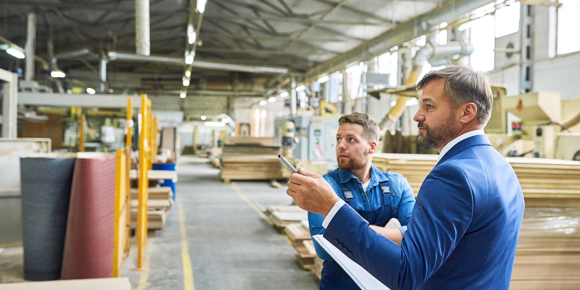 Der Mann in Anzug und weißem Hemd zeigt mit einem Stift selbstbewusst nach links. Ein Arbeiter in blauer Arbeitskleidung blickt entschlossen in die gleiche Richtung. Beide stehen in einer Fabrikhalle. © Adobe Stock, Seventyfour