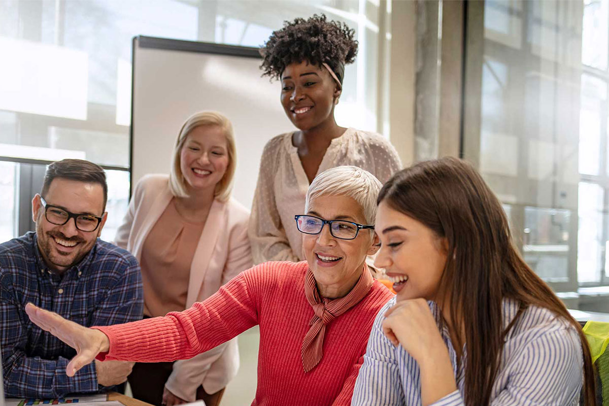 Mitarbeiter und Azubis sind um einen Arbeitsplatz versammelt und lachen. © Adobe Stock, Dragana Gordic