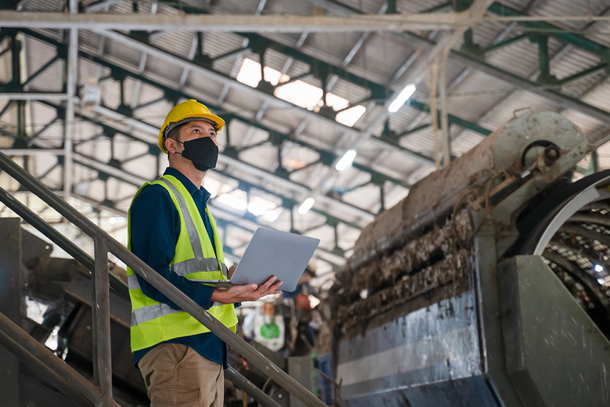 Sicherheitsbeauftragte der betriebsärztlichen und sicherheitstechnischen Betreuung mit Laptop, Maske und Warnweste inspizieren Produktionsanlage in Industriehalle. © Adobe Stock, Attasit