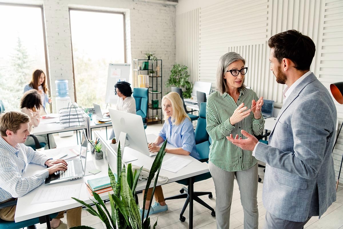 Ältere und jüngere Mitarbeitende arbeiten in einem modernen Büro, während eine ältere Angestellte mit einem jüngeren Mitarbeiter diskutiert. © Adobe Stock, deagreez