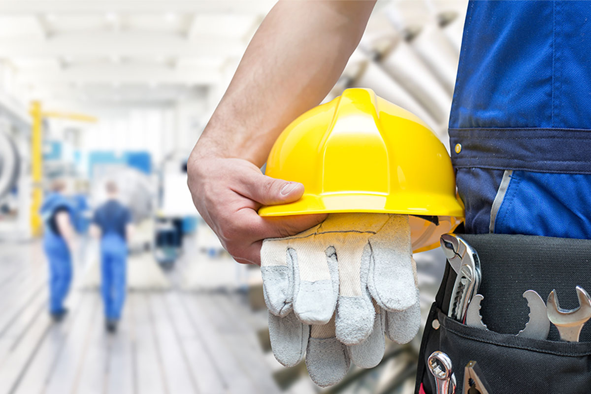 Arbeiterhand hält gelben Schutzhelm und Handschuhe, Werkzeuge ragen aus der Gürteltasche. © Adobe Stock, Industrieblick