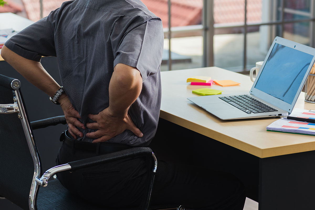 Mann sitzt Rückenschmerzen an seinem Arbeitsplatz, welcher nicht den Technischen Regeln entspricht.